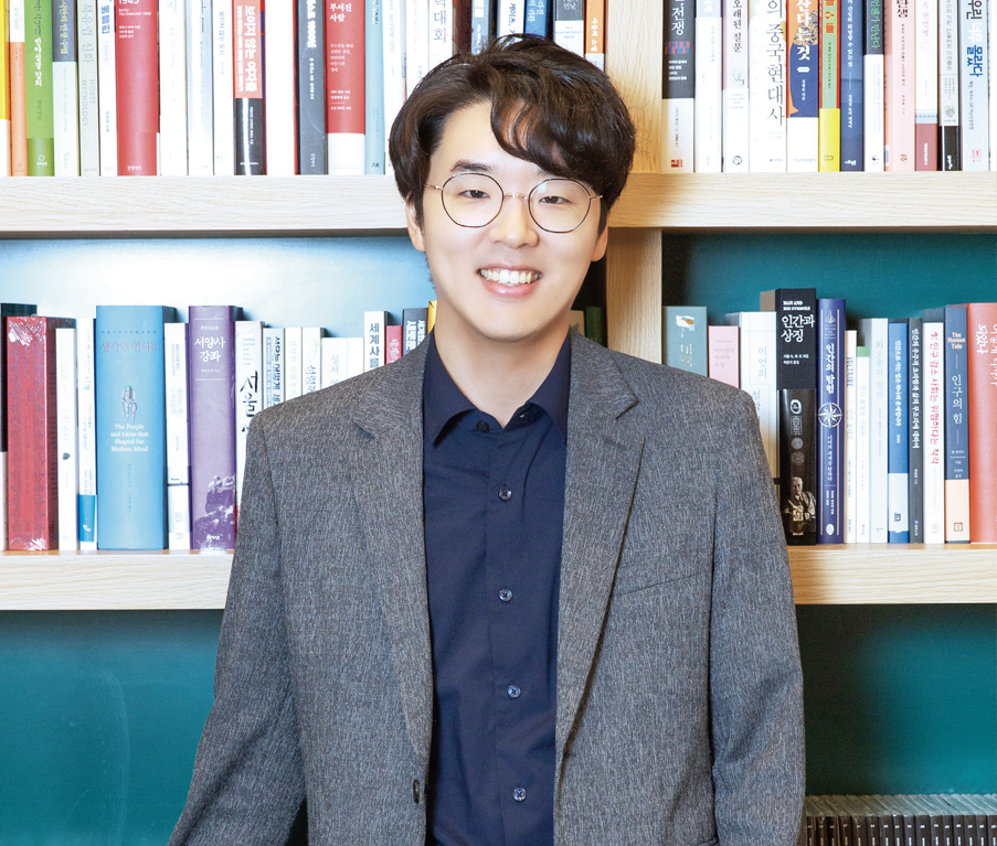 책장을 배경으로 정면을 보고 서 있는 정지우 작가